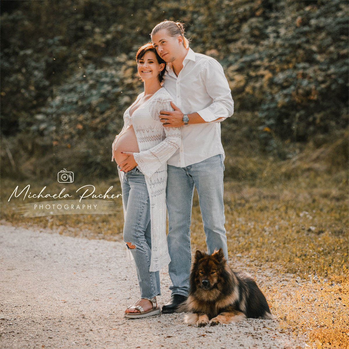 Schwangere Frau steht mit ihrem Partner auf einem Waldweg, beide lächeln entspannt in die Ferne; der Hund der Familie liegt ruhig zu ihren Füßen. Warmes Abendlicht sorgt für eine natürliche, harmonische Stimmung.